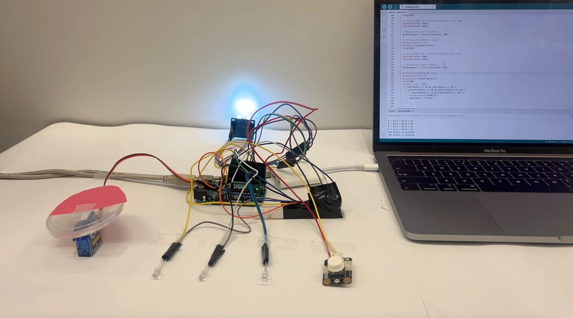The electronics on the bench: a colour sensor, a servo-driven tray, three LEDs and a button on an Arduino Uno, with the readings in the serial monitor.